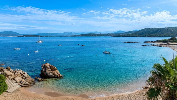 A coastal beach with clear blue water, boats, and rocky formations.