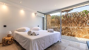 A modern bedroom with a large bed, a wooden side table, and a view of a natural landscape through a glass door.