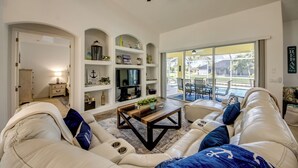 A spacious living room with a white sofa set, a coffee table, and a TV. There are shelves with decorative items and a view of the outside through a glass door. - Villa Latitude Adjustment - Spacious 4 BR/ 3 bath waterfront Villa with heated pool and spa, kayaks and bikes (Cape Coral)