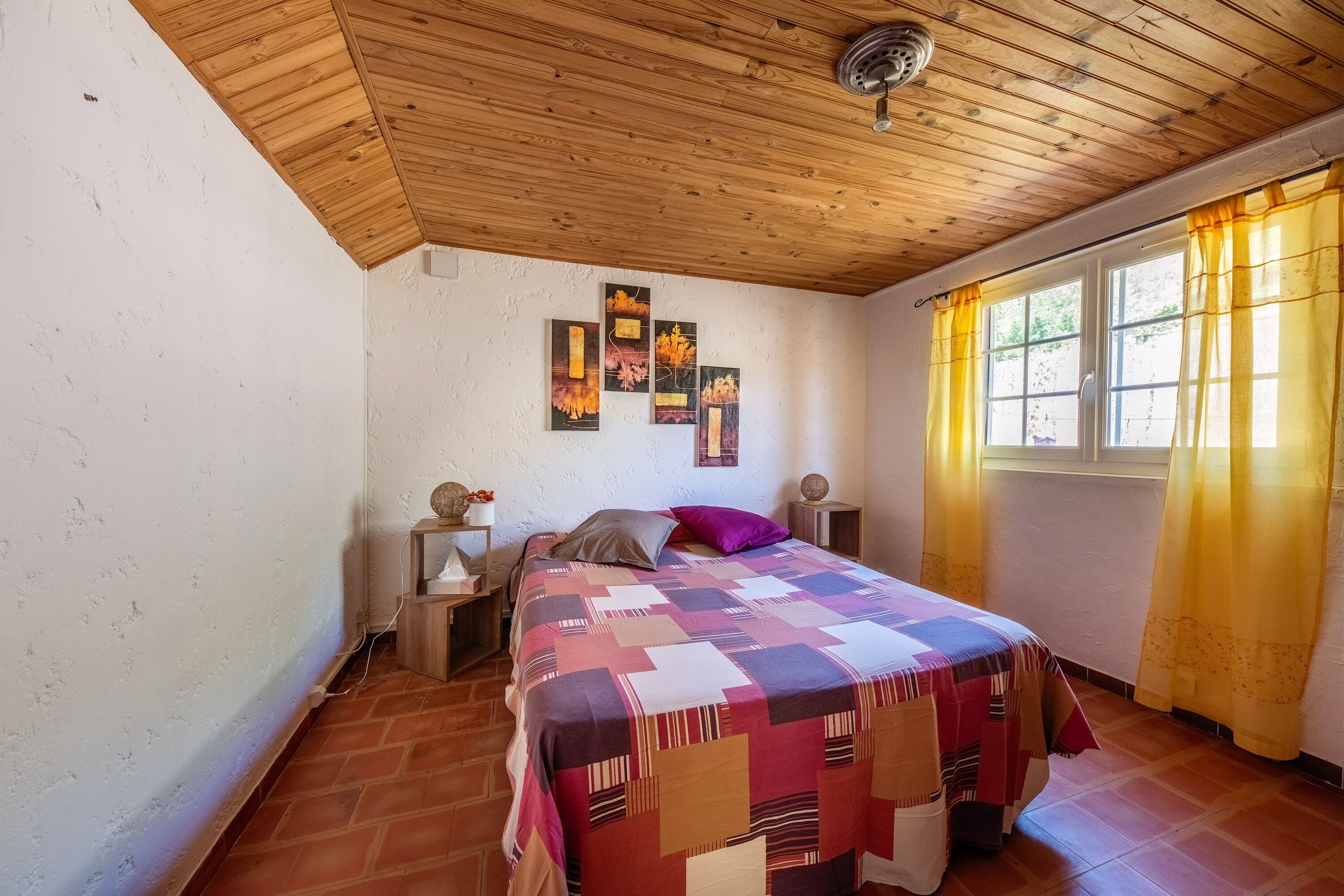 A bedroom with a wooden ceiling, a bed with a patterned bedspread, a nightstand with a lamp, and a window with yellow curtains.