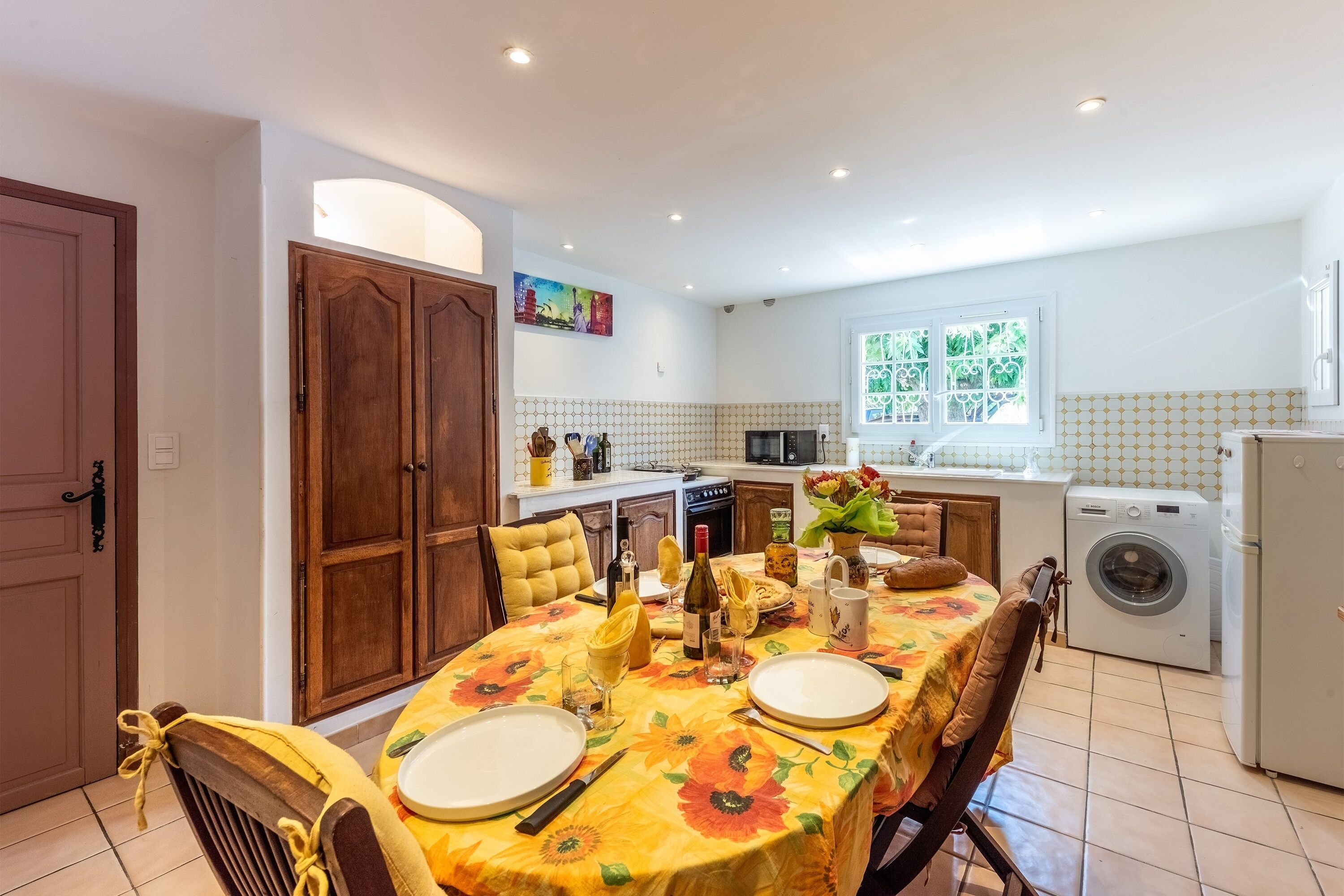 A dining area with a table set for two, a kitchen in the background, and a washing machine to the right.