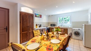 A dining area with a table set for two, a kitchen in the background, and a washing machine to the right. - La Bouscarle - Jardin et Piscine Partagée (Châteaudouble)