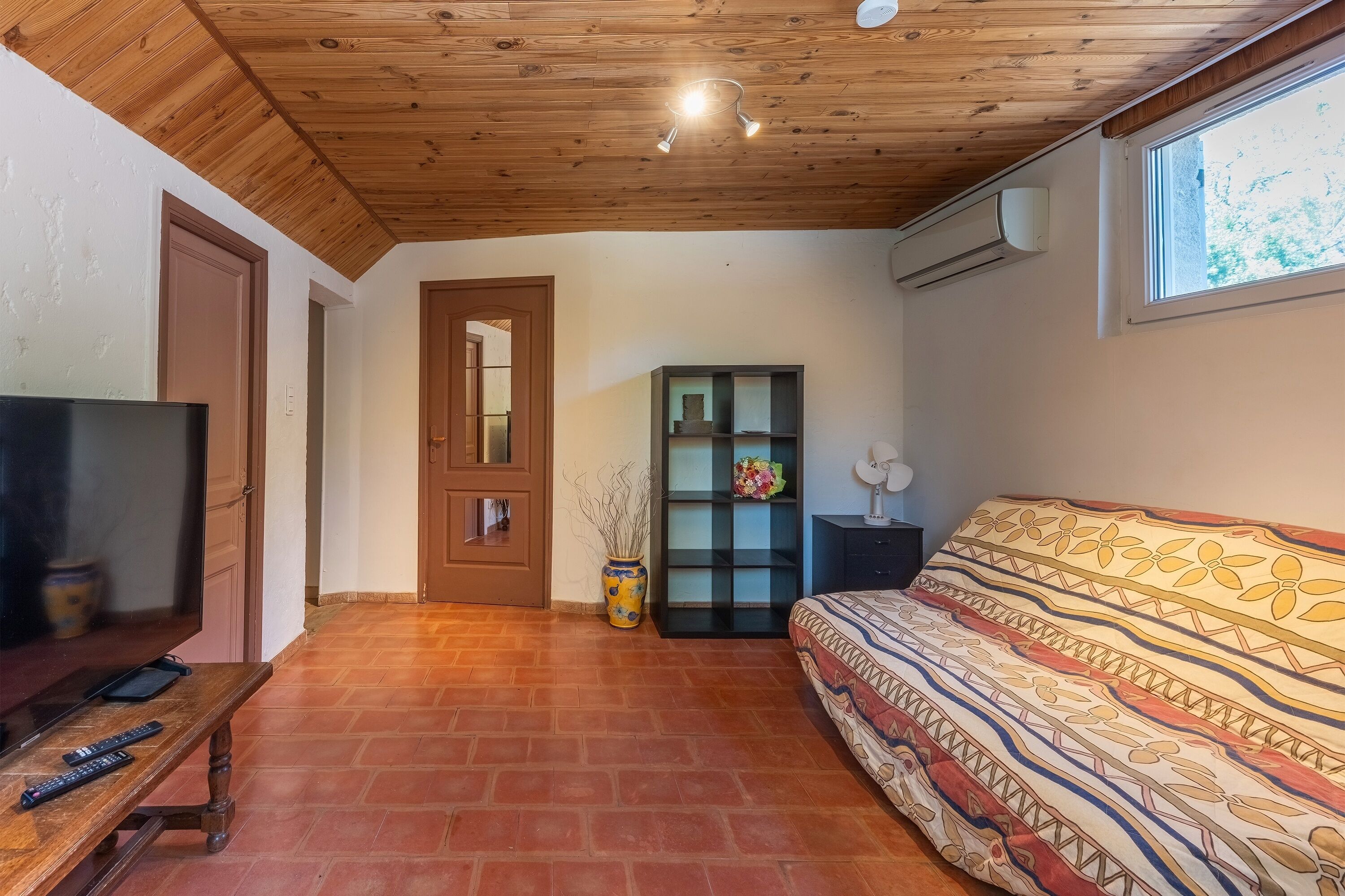 A room with a wooden ceiling, a television, a sofa, a bookshelf, and a window.