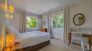 Ein Schlafzimmer mit Bett, Nachttischlampe, Schminktisch, Spiegel und fensterbepflanzten Fenstern.