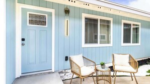 A light blue building with a front door, two windows, and a small outdoor seating area with wicker chairs and a table.