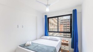 A bedroom with a bed, a nightstand, a window with blue curtains, and a ceiling fan. - Beachfront Building With Pool - CPK (Ilhéus)