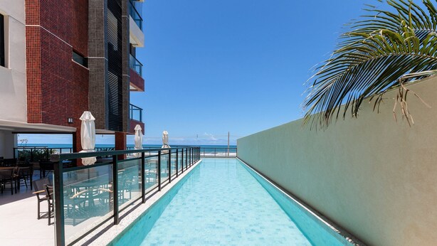 A pool area with a clear view of the ocean, a palm tree, and a building with red and grey tiles. - Beachfront Building With Pool - CPK (Ilhéus)