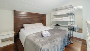 Een slaapkamer met een houten hoofdbord, een grijze dekbedovertrek, een witte nachtkastje en een metalen plank voor opgevouwen handdoeken.