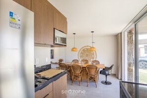 A modern kitchen with wooden cabinets, a black countertop, and a dining area with wicker chairs and a glass table. - NLU - Full-featured Condominium at Marceneiro Beach (Passo de Camaragibe)