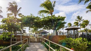 A wooden boardwalk leading to a building with a thatched roof, surrounded by palm trees and lush greenery. - NLU - Full-featured Condominium at Marceneiro Beach (Passo de Camaragibe)