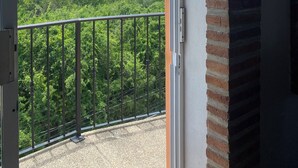 A balcony with a metal railing, a brick wall, and a glass door leading to a view of a green landscape and a clear sky. - Casitas Las Palmas (Punta de Mita)