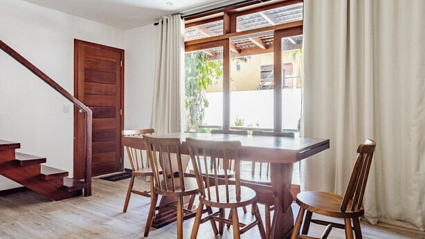 A dining area with a wooden table and chairs, a wooden staircase, and a view through a window with curtains.
