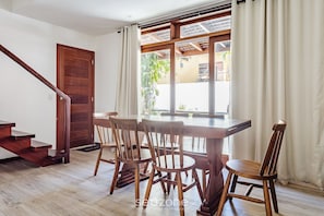 A dining area with a wooden table and chairs, a staircase, and a view through a window with curtains. - House With Pool in the Condominium Cvb0144 (Maraú)
