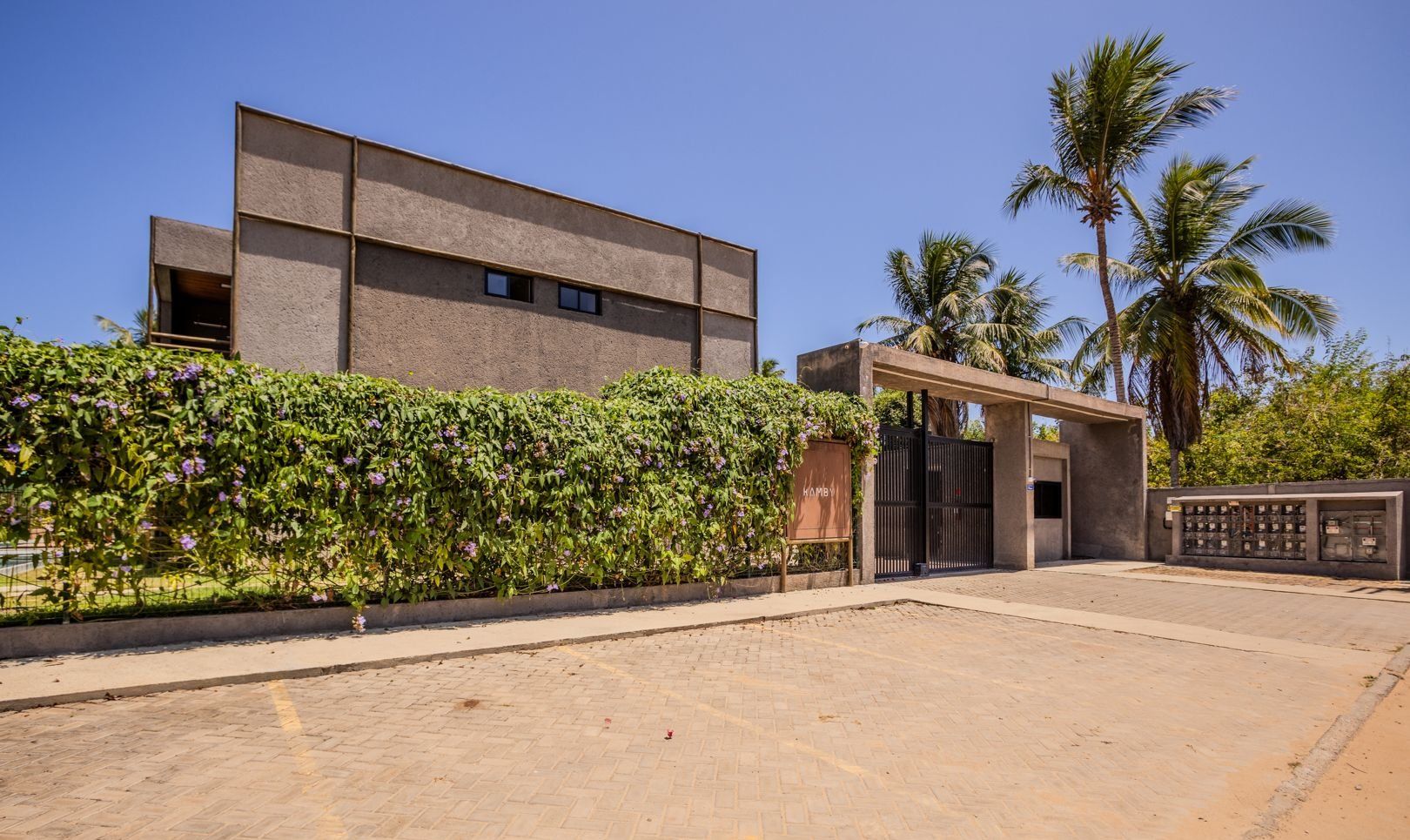 A modern building with a flat roof, a gated entrance, and a landscaped area with a hedge and palm trees.