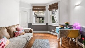 Um quarto aconchegante com um sofá de couro marrom, uma pequena mesa de jantar com cadeiras amarelas e uma janela com vista para uma escadaria de tijolos.