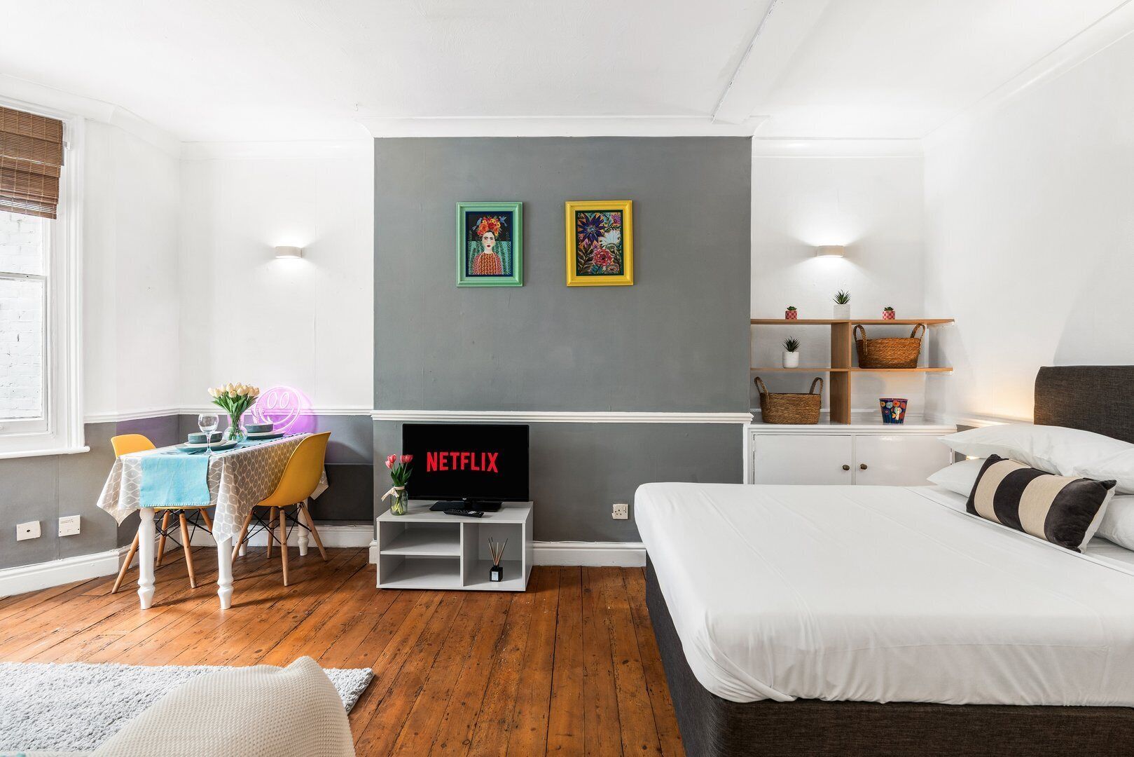 Ein modernes Schlafzimmer mit Bett, Fernseher, Essbereich mit Tisch und Stühlen sowie einer grauen Akzentwand mit zwei gerahmten Bildern.