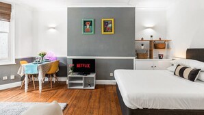 Quarto moderno com cama, televisão, área de refeições com mesa e cadeiras, e uma parede cinza com duas molduras.