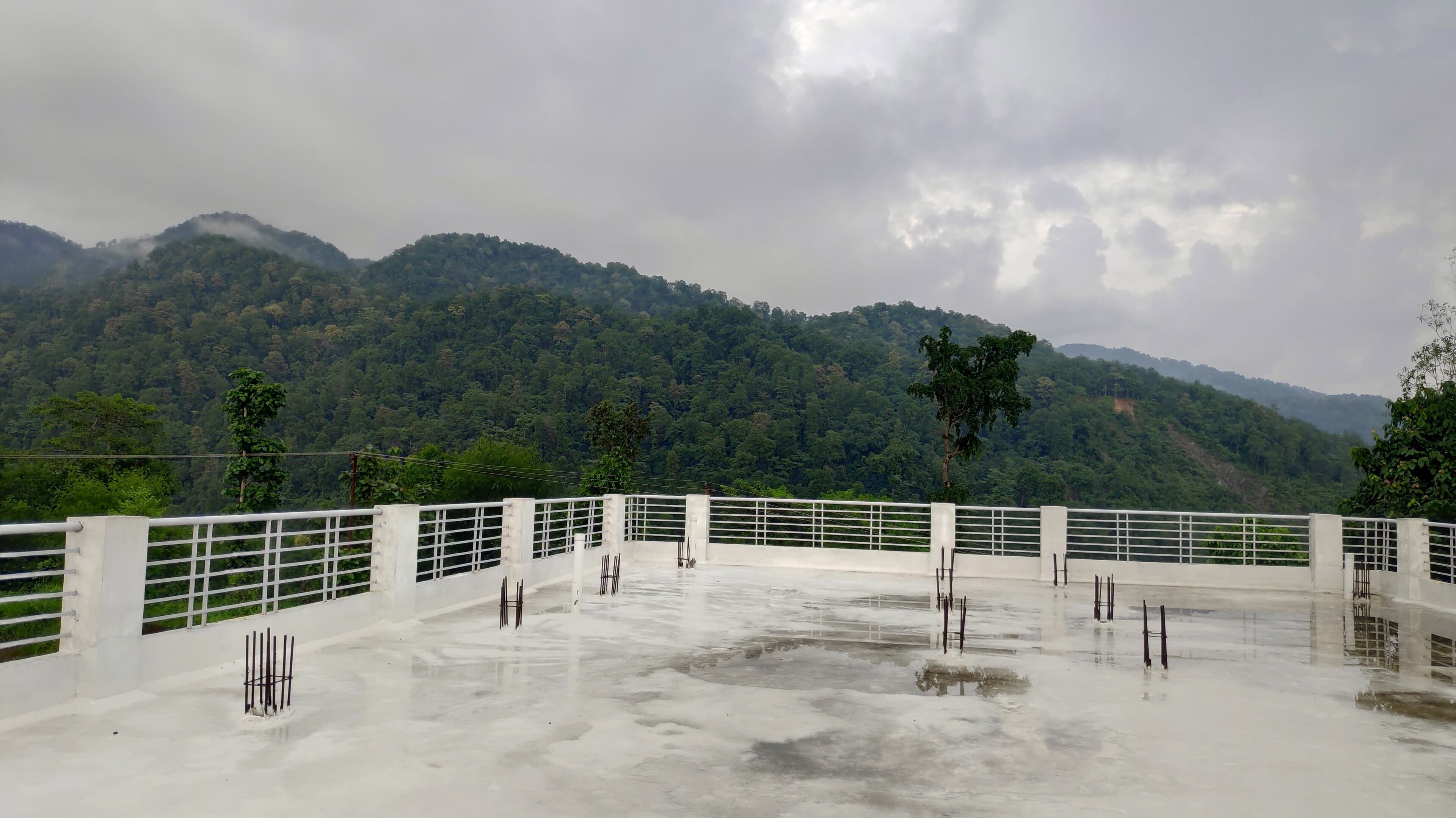 A concrete terrace with white railings, surrounded by lush green hills under a cloudy sky.