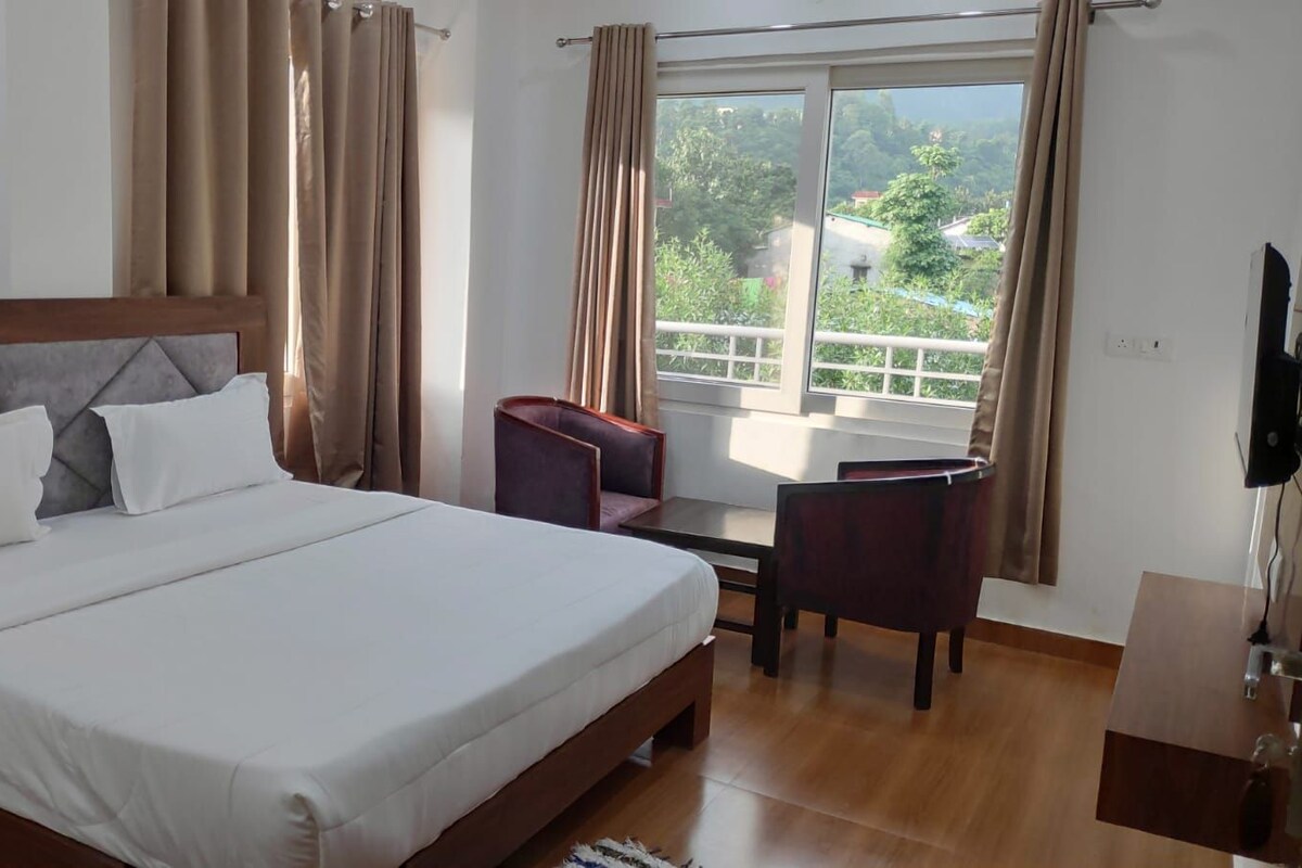 A hotel room with a large bed, a desk, and a chair. There is a window with curtains and a view of greenery outside.