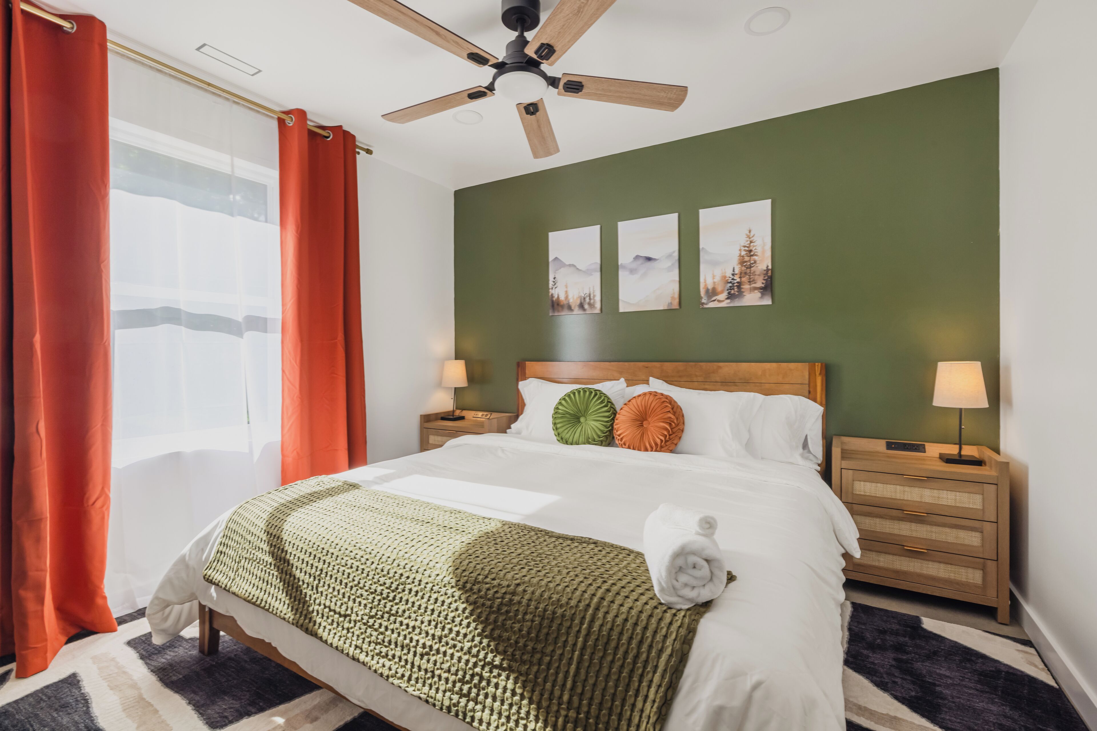 Een net opgemaakt bed met een groene deken, een houten hoofdbord en twee kussens. Er zijn twee nachtkastjes met lampen, een plafondventilator en een raam met rode gordijnen.
