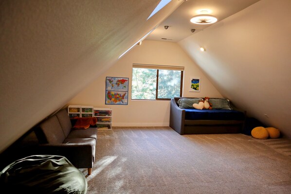 A cozy attic bedroom with a sloped ceiling, a large window, a bed with pillows, a small couch, and storage boxes. - Forest Retreat at Canyon Loop (Flagstaff)