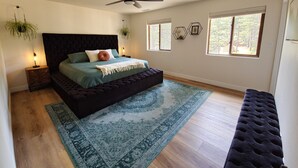 A bedroom with a dark headboard, a bed with multiple pillows, a blue patterned rug, a ceiling fan, and two windows with a view of trees. - Forest Retreat at Canyon Loop (Flagstaff)