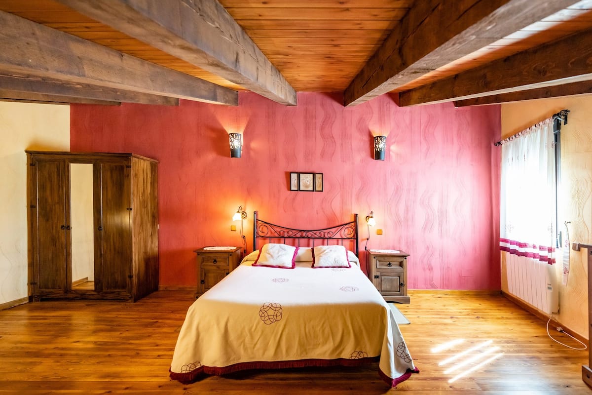 Une chambre avec un plafond en bois, un lit recouvert d’une courtepointe blanche, deux tables de chevet, une armoire en bois et une fenêtre avec des rideaux.