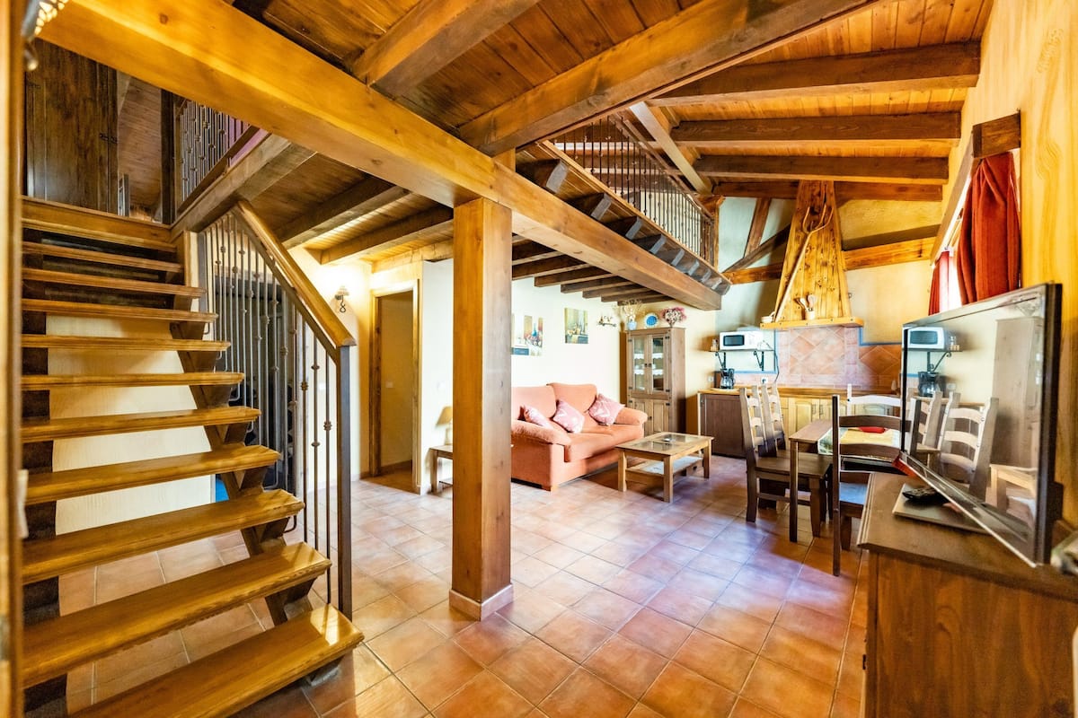 Un escalier en bois mène à une chambre située en mezzanine, un salon avec un canapé et un coin repas avec des chaises et une table.