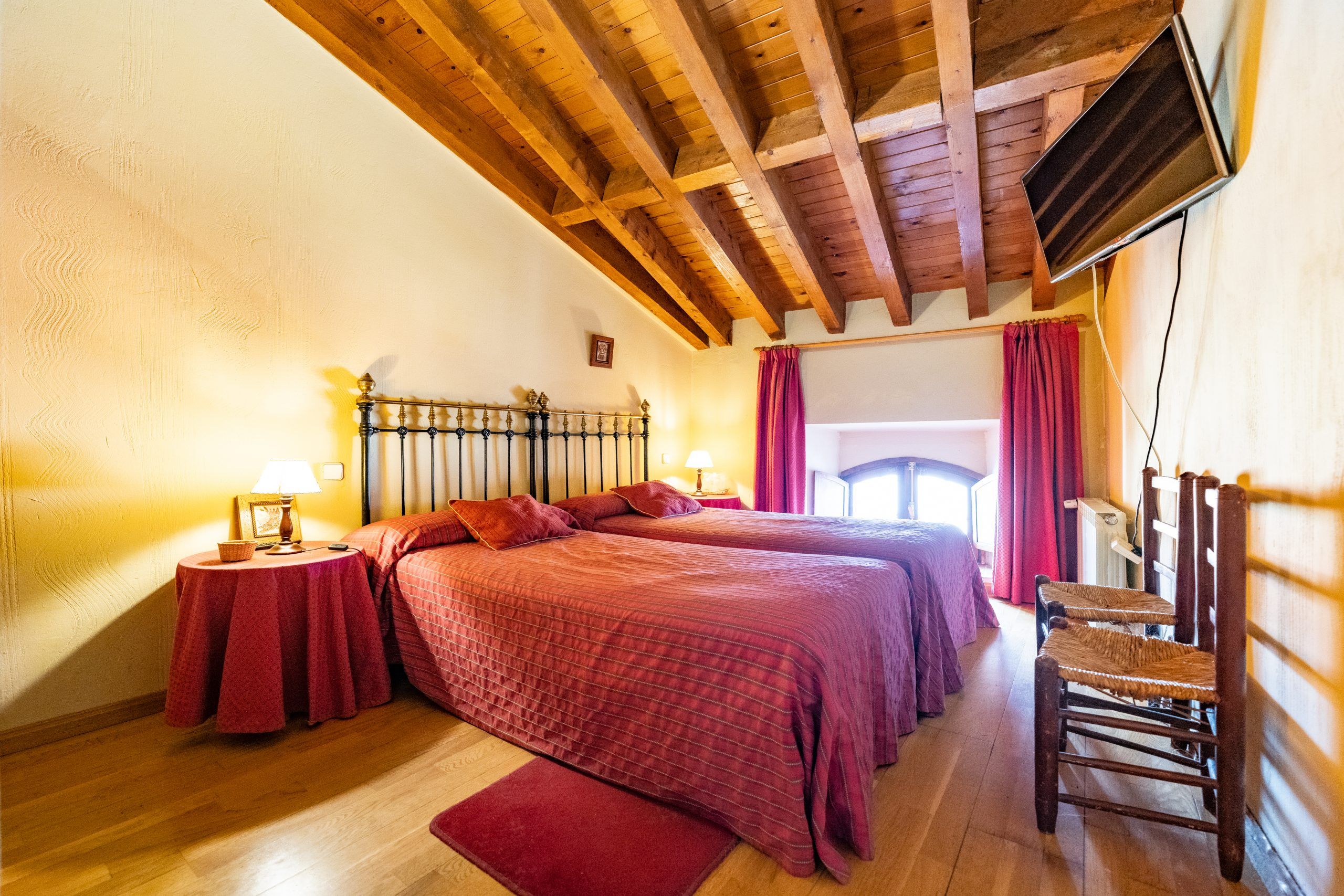 A bedroom with two beds, wooden beams on the ceiling, a TV mounted on the wall, and a chair.