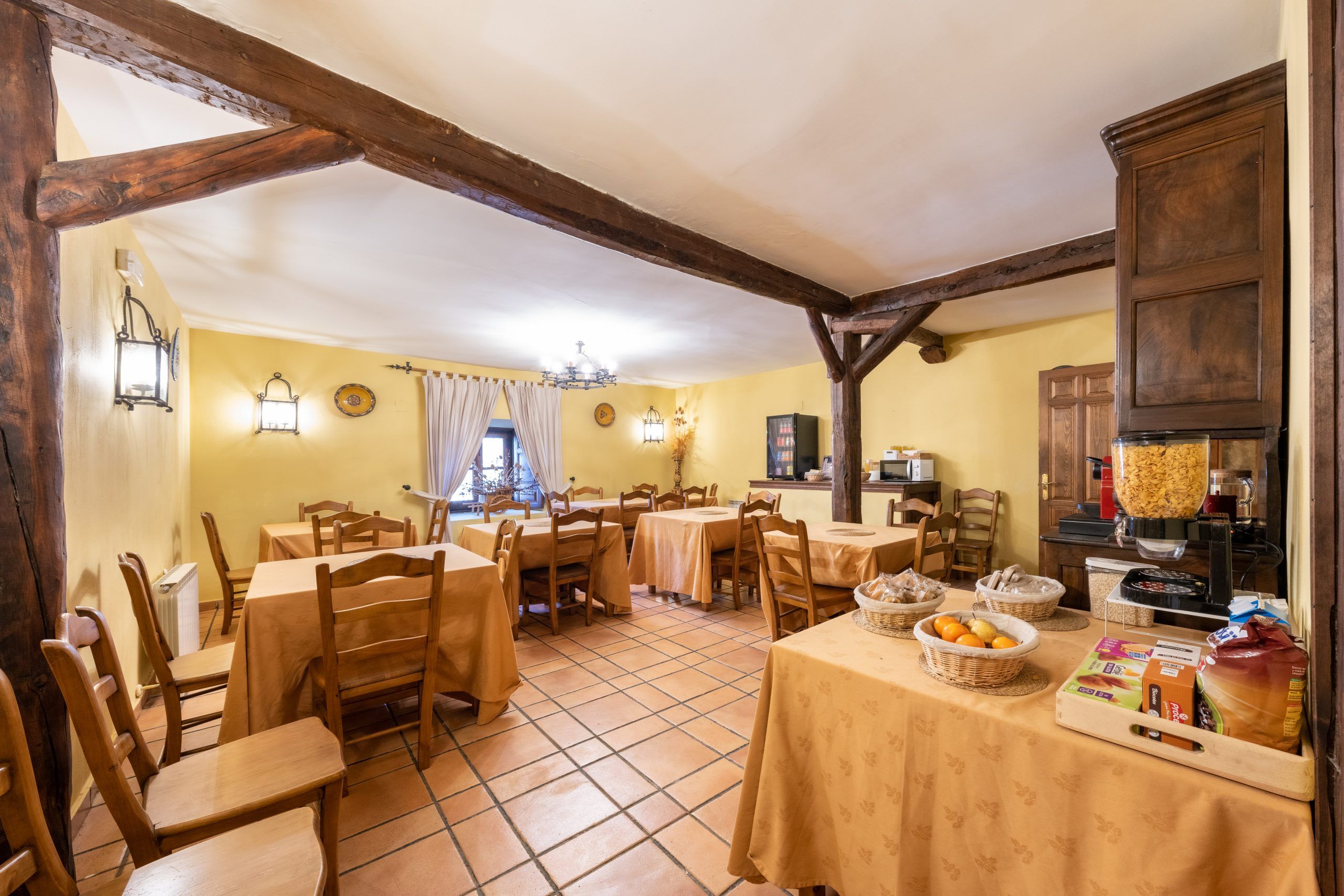 Un comedor con mesas y sillas de madera, una zona de cocina con una máquina para pan y un suelo de baldosas.