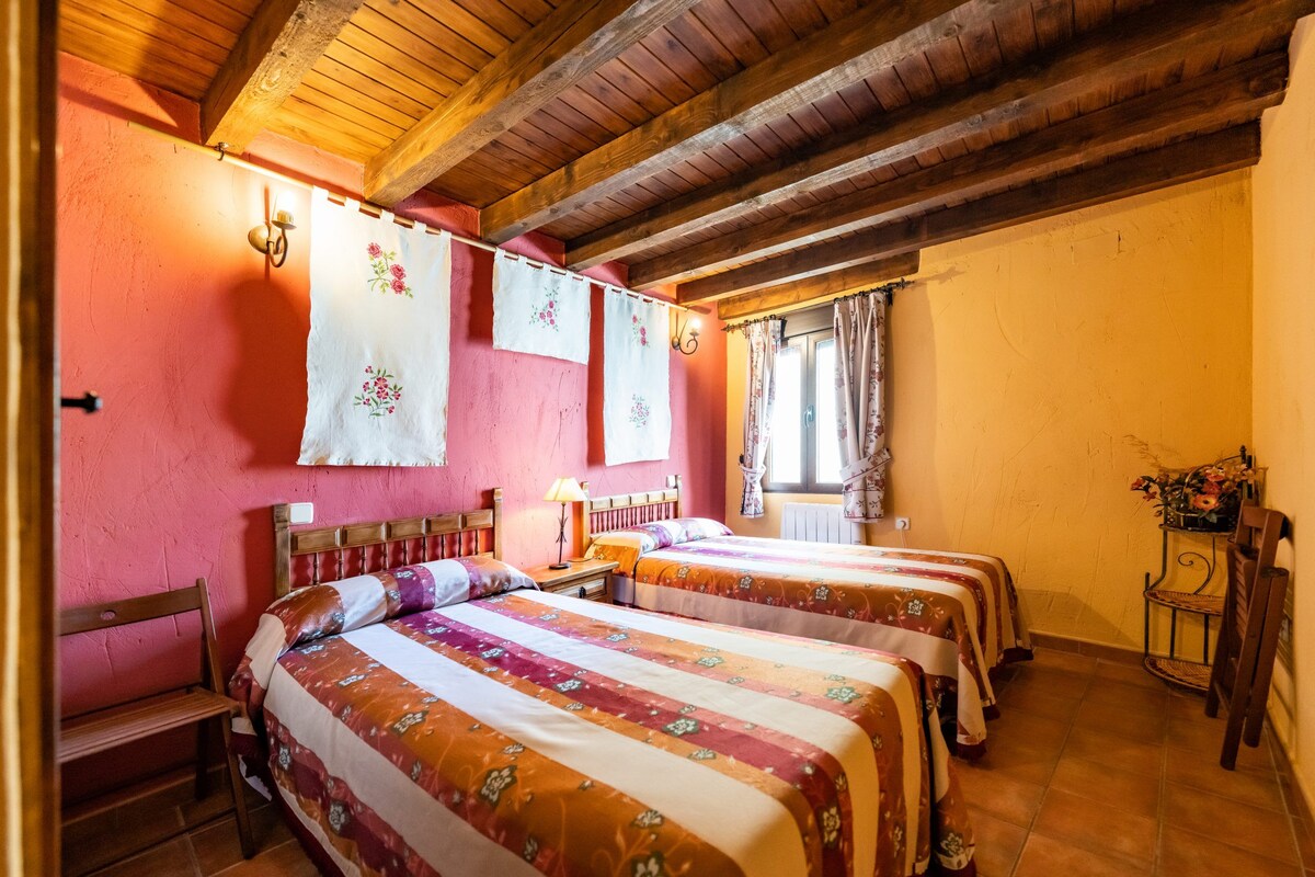 A room with two beds, wooden beams on the ceiling, a bench, and a small table with a lamp.