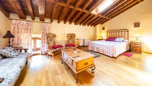 A cozy room with a wooden ceiling, a bed with a white and pink bedspread, a wooden chest of drawers, a sofa with floral upholstery, and two armchairs with red cushions.