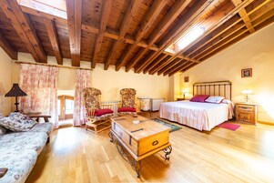 Una habitación acogedora con techo de madera, una cama con colcha blanca y rosa, un aparador de madera, un sofá con tapicería floral y dos sillones con cojines rojos.