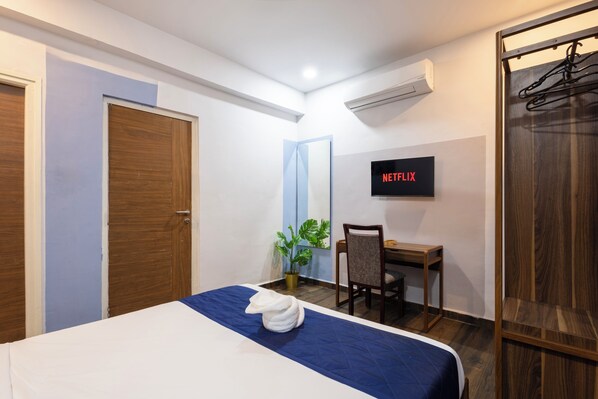 A hotel room with a bed, a desk, a chair, a potted plant, and a wall-mounted TV displaying 'NETFLIX'.