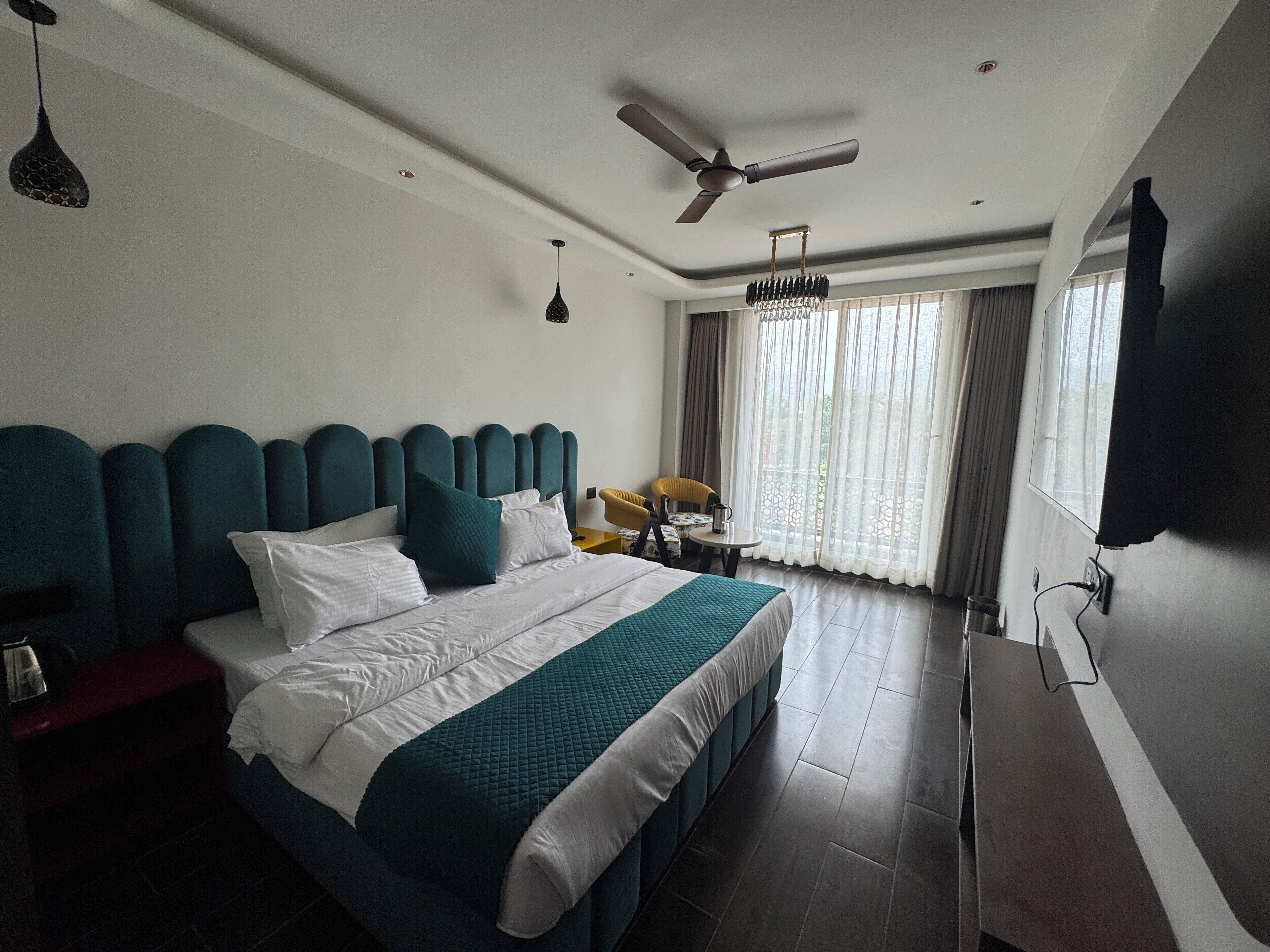 A hotel room with a bed, a ceiling fan, a table with chairs, and a view of the outdoors through large windows.