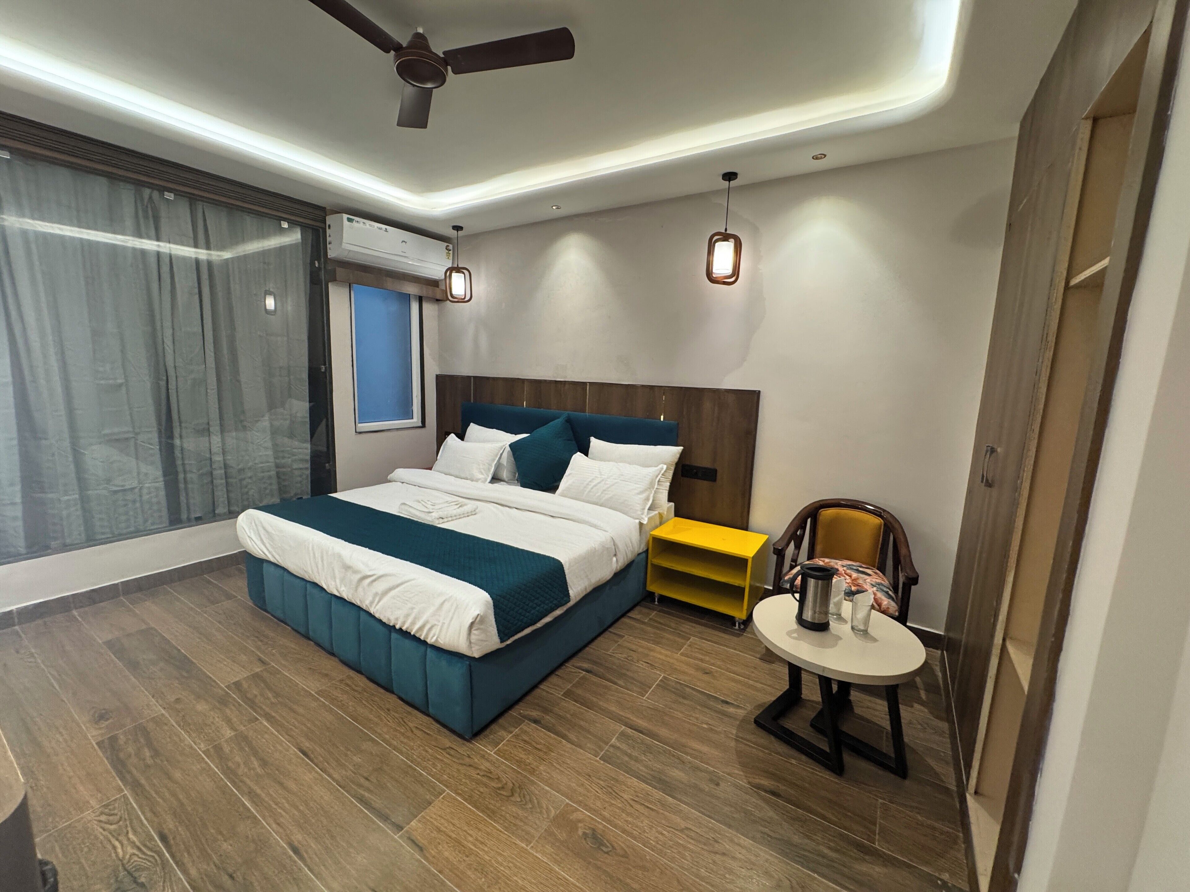 A modern bedroom with a large bed, a ceiling fan, a small table with a chair, and a window with curtains.