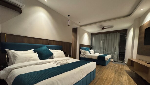 A modern hotel room with two beds, a large window with curtains, a ceiling fan, and a flat-screen TV. - Hotel Ganga Aamantran (Haridwar)