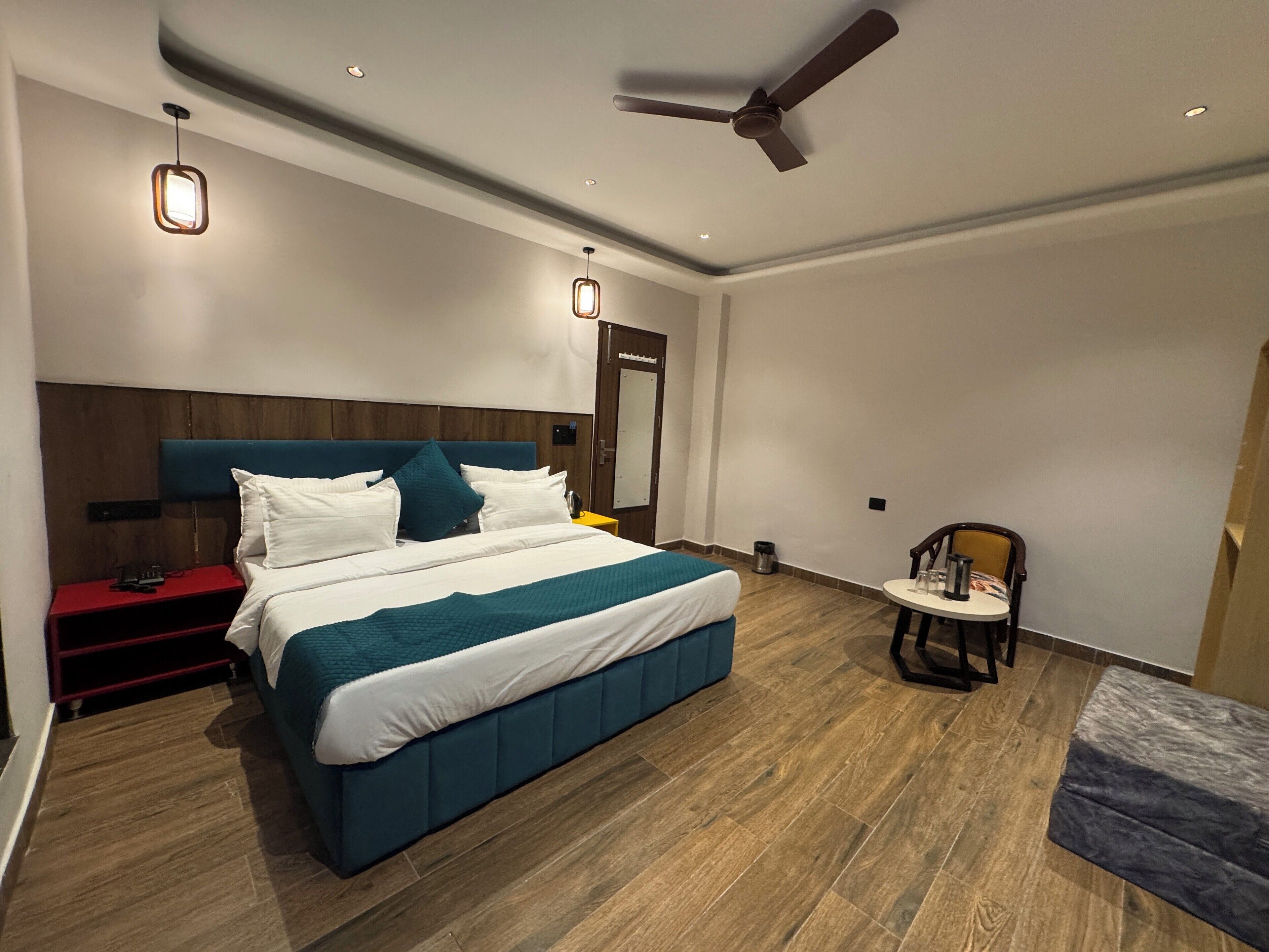 A modern hotel room with a large bed, a ceiling fan, a small table with chairs, and a red nightstand.