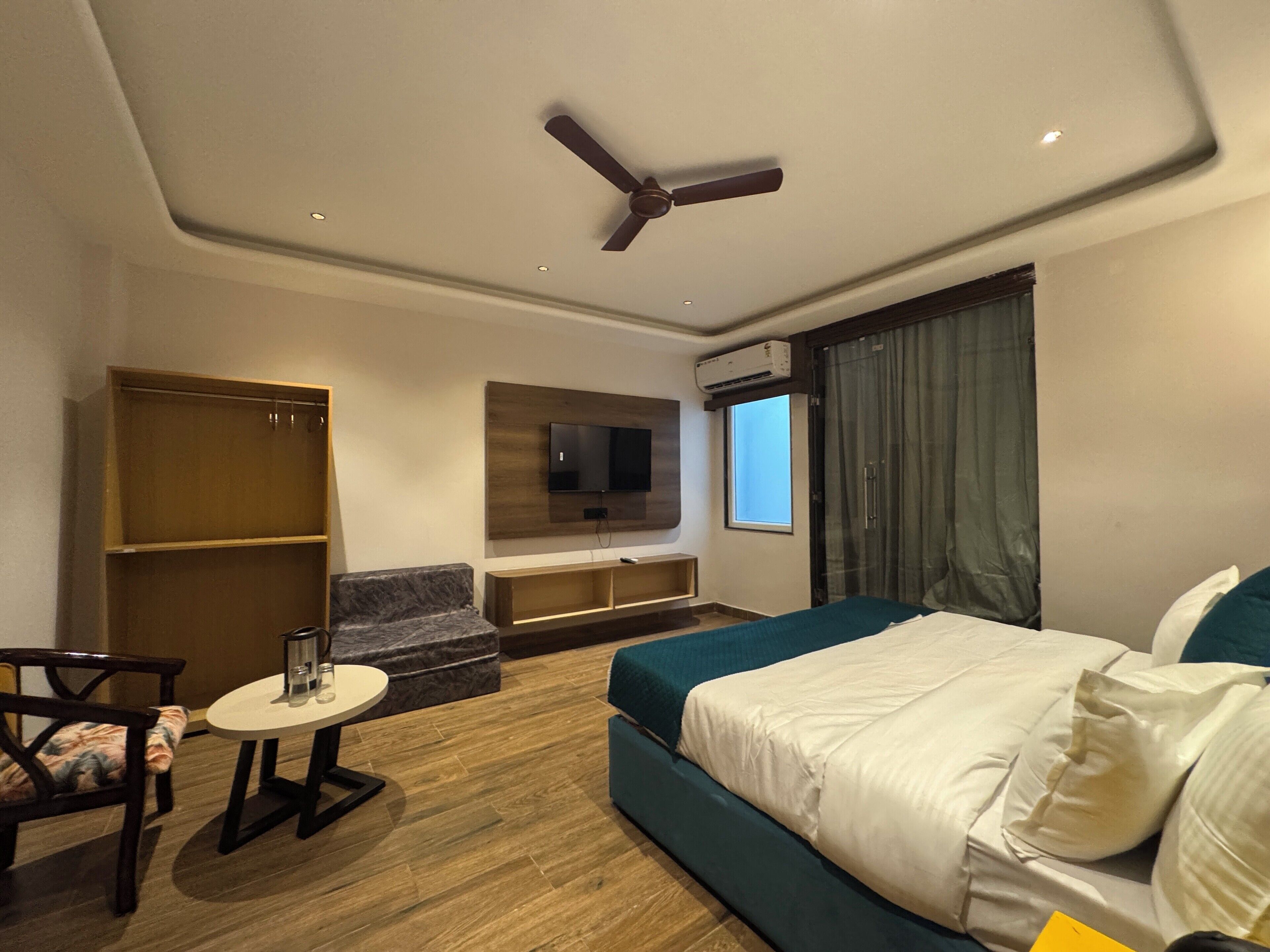 A hotel room with a bed, a TV mounted on the wall, a ceiling fan, a window with curtains, and a seating area with a table and chairs.