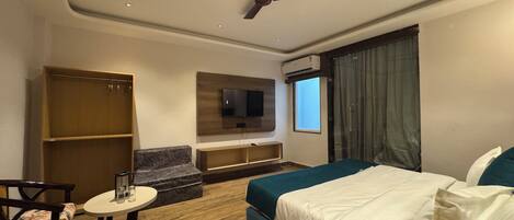 Une chambre d’hôtel comprenant un lit, un téléviseur fixé au mur, un ventilateur de plafond, une fenêtre avec des rideaux et un coin salon avec une table et des chaises.