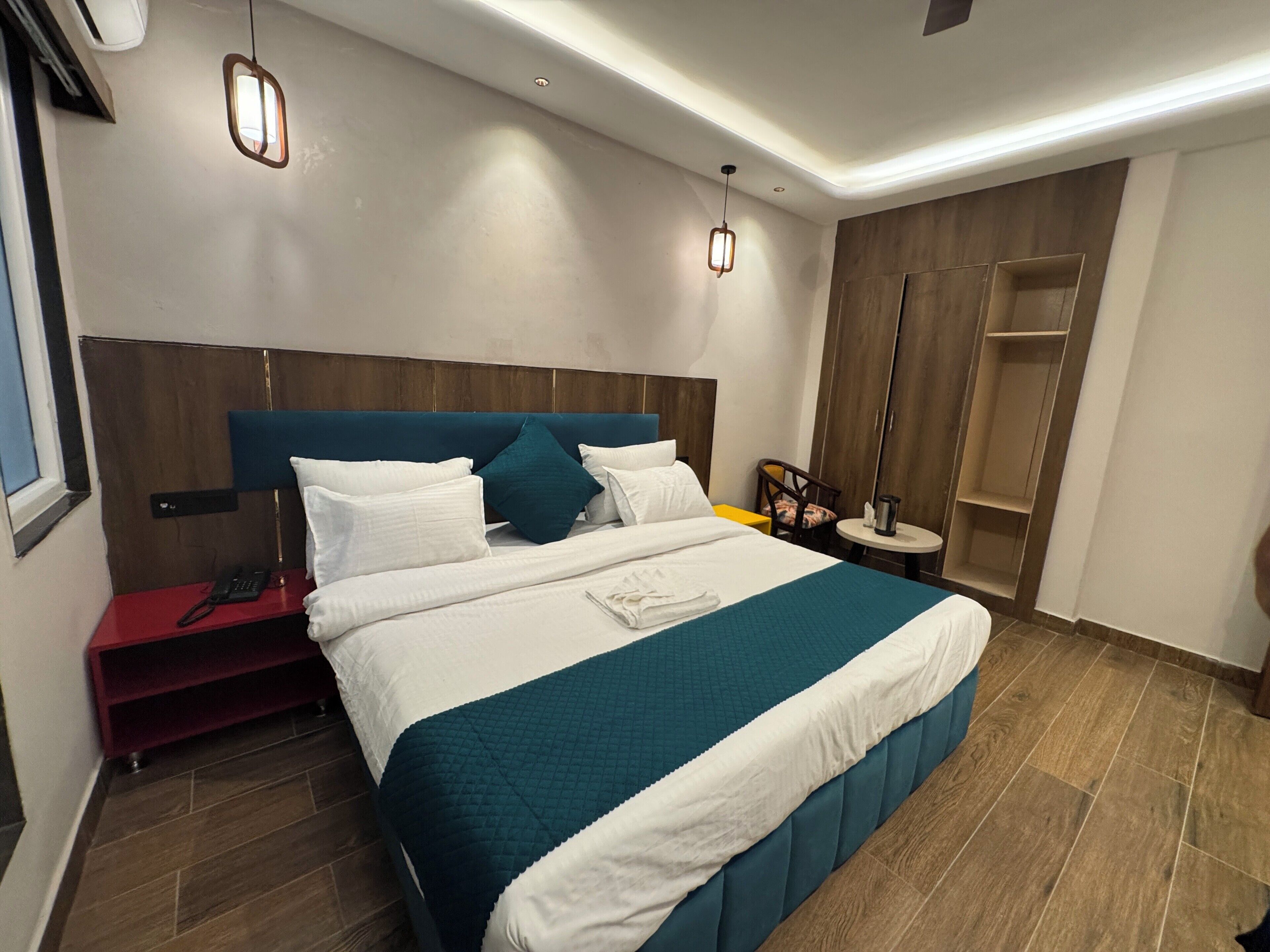 A modern hotel room with a large bed, a bedside table, a red nightstand, a wooden wardrobe, and a small round table with a lamp.