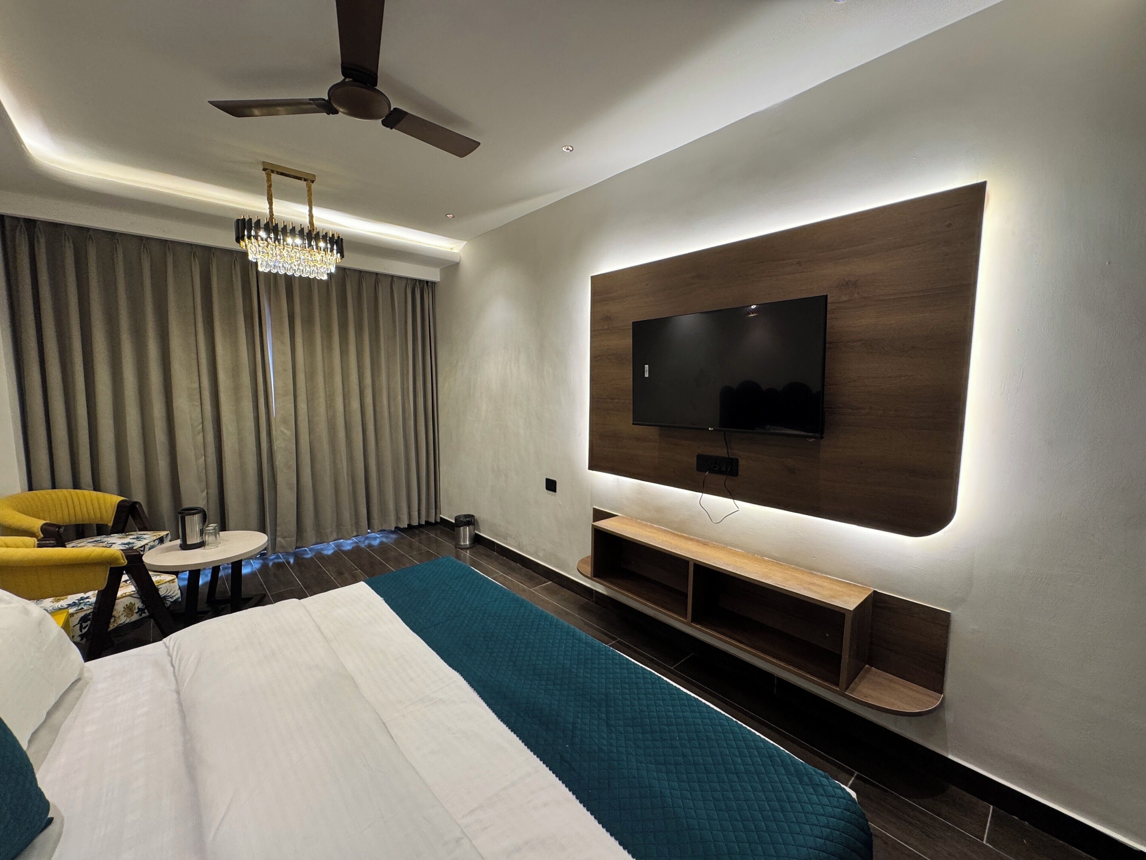 Una habitación de hotel moderna con una cama grande, un televisor de pantalla plana montado en un panel de madera, un ventilador de techo y una araña.
