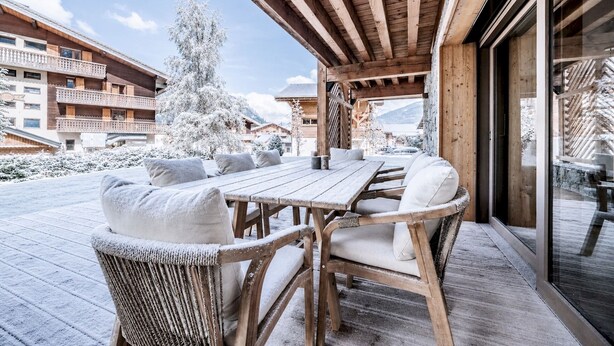 A wooden deck with a table and chairs, surrounded by snow-covered trees and buildings.