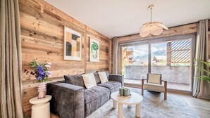 A modern living room with a wooden wall, a grey sofa, a round coffee table, and a large window with a view of a mountain.