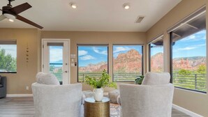 A living room with two grey armchairs, a round coffee table with a white vase and green plant, and a patterned rug. Large windows offer a view of red rock formations and greenery. - Mid-century Gem w/ Views, Hot Tub & Sunroom Lounge Red Rock Haven by Avantstay (Sedona)