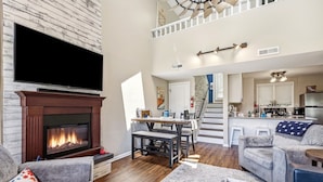 A spacious living room with a fireplace, a dining area, and a kitchen with a staircase leading to an upper level.