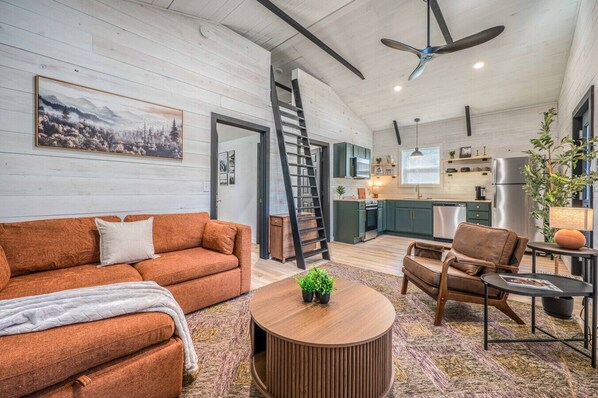 Una sala de estar moderna con sofá, sillón y mesa de centro, una cocina al fondo y una escalera que sube a otro nivel.