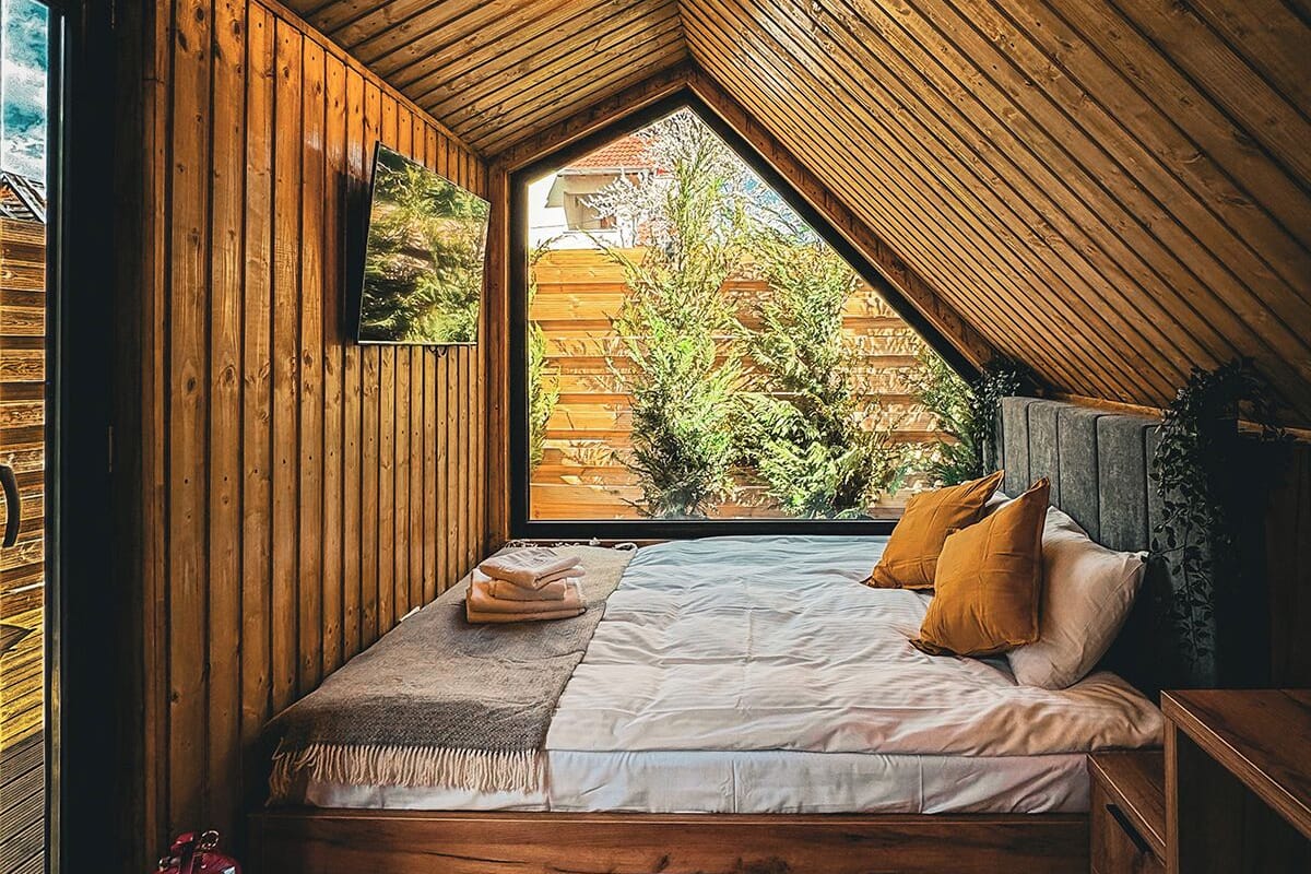 A cozy attic bedroom with wooden walls, a sloped ceiling, a bed with pillows, a small table, and a window overlooking greenery.