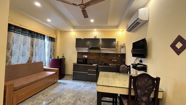 A compact living space with a sofa, dining table, chairs, a kitchen area with cabinets and appliances, and a ceiling fan. - Kedar inn (Rishikesh)