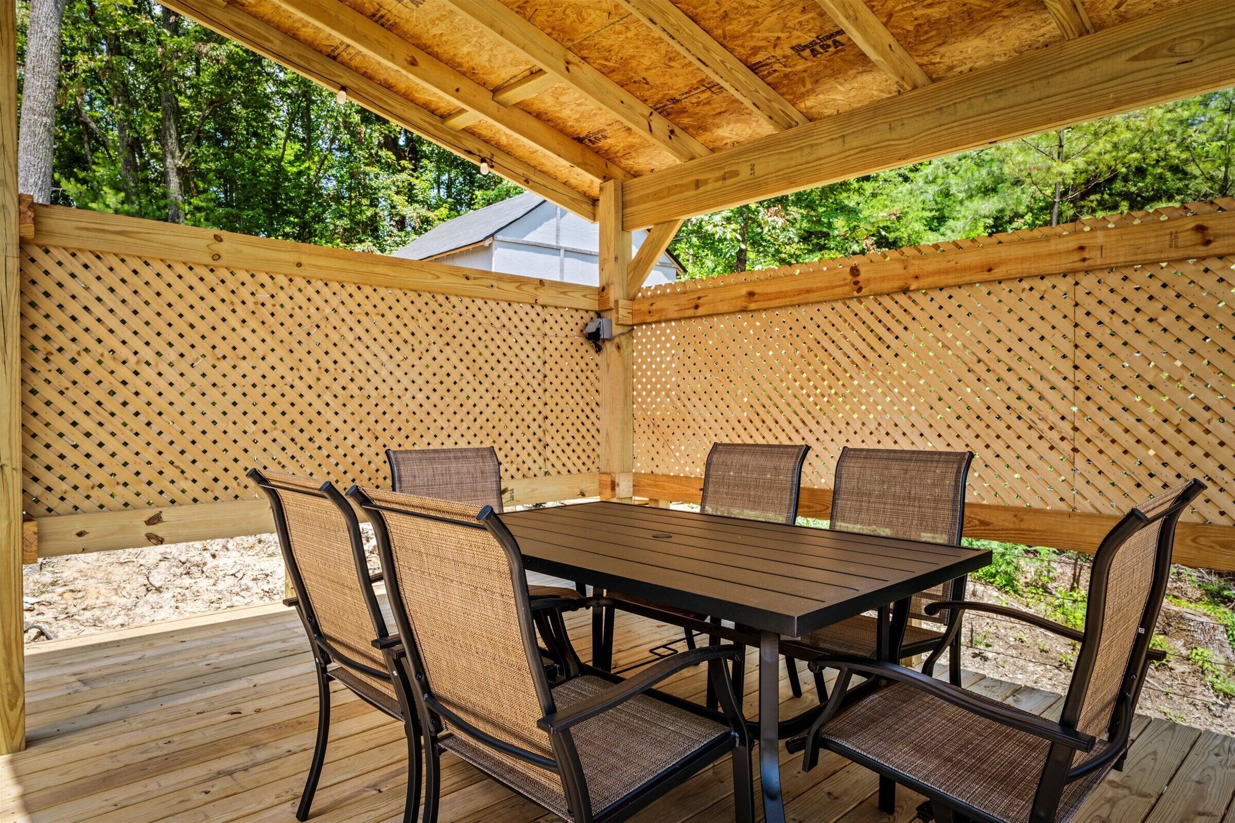 A covered outdoor patio with a wooden table and chairs, surrounded by a wooden fence with a lattice design.